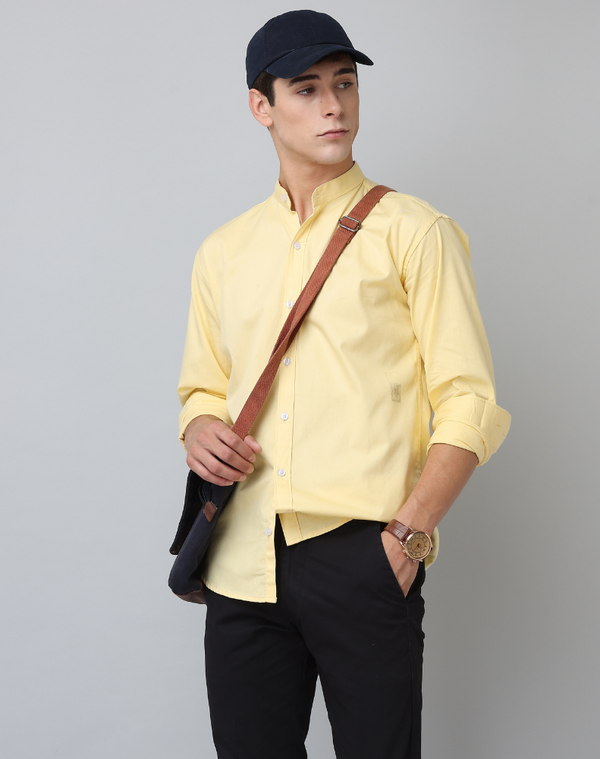 Man wearing a yellow shirt, black pants, and a black cap with a brown strap over his shoulder on a gray background.