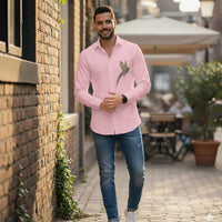 Man wearing a pink shirt with a bird design and blue jeans standing against a brick wall.