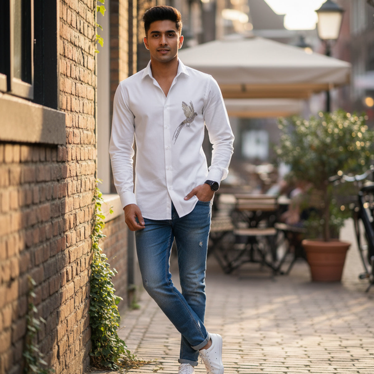 Man in a white shirt with a bird design standing against a brick wall in an outdoor setting.